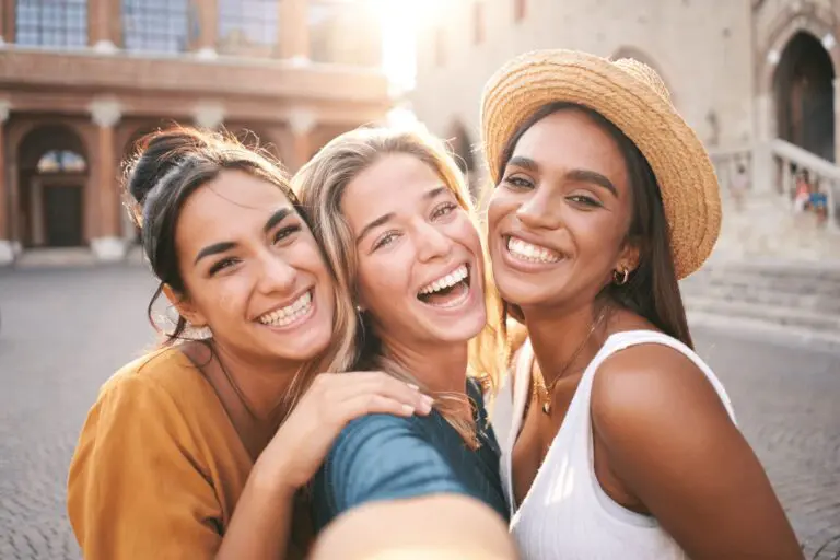 Three young women smile and take a selfie outdoors in a sunlit, urban setting with historic buildings, close to Smile City Orthodontics in Sunnyvale, TX.