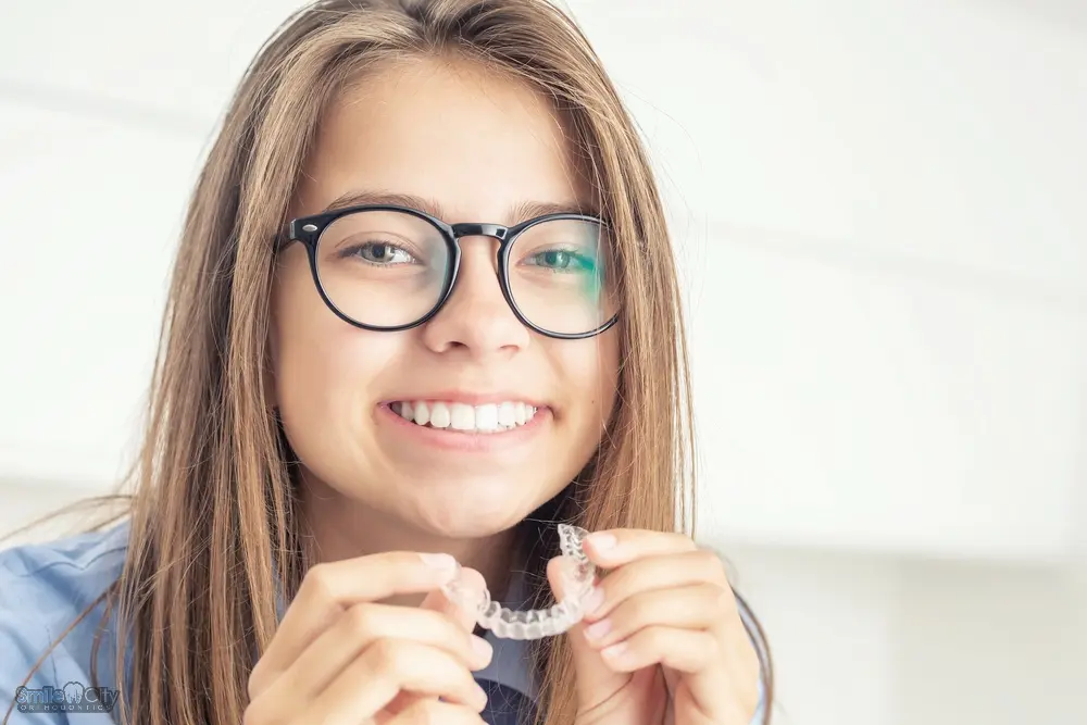 A young woman with glasses holding a clear aligner and smiling, showing an alternative method for how to fix overbite in Sunnyvale, TX.