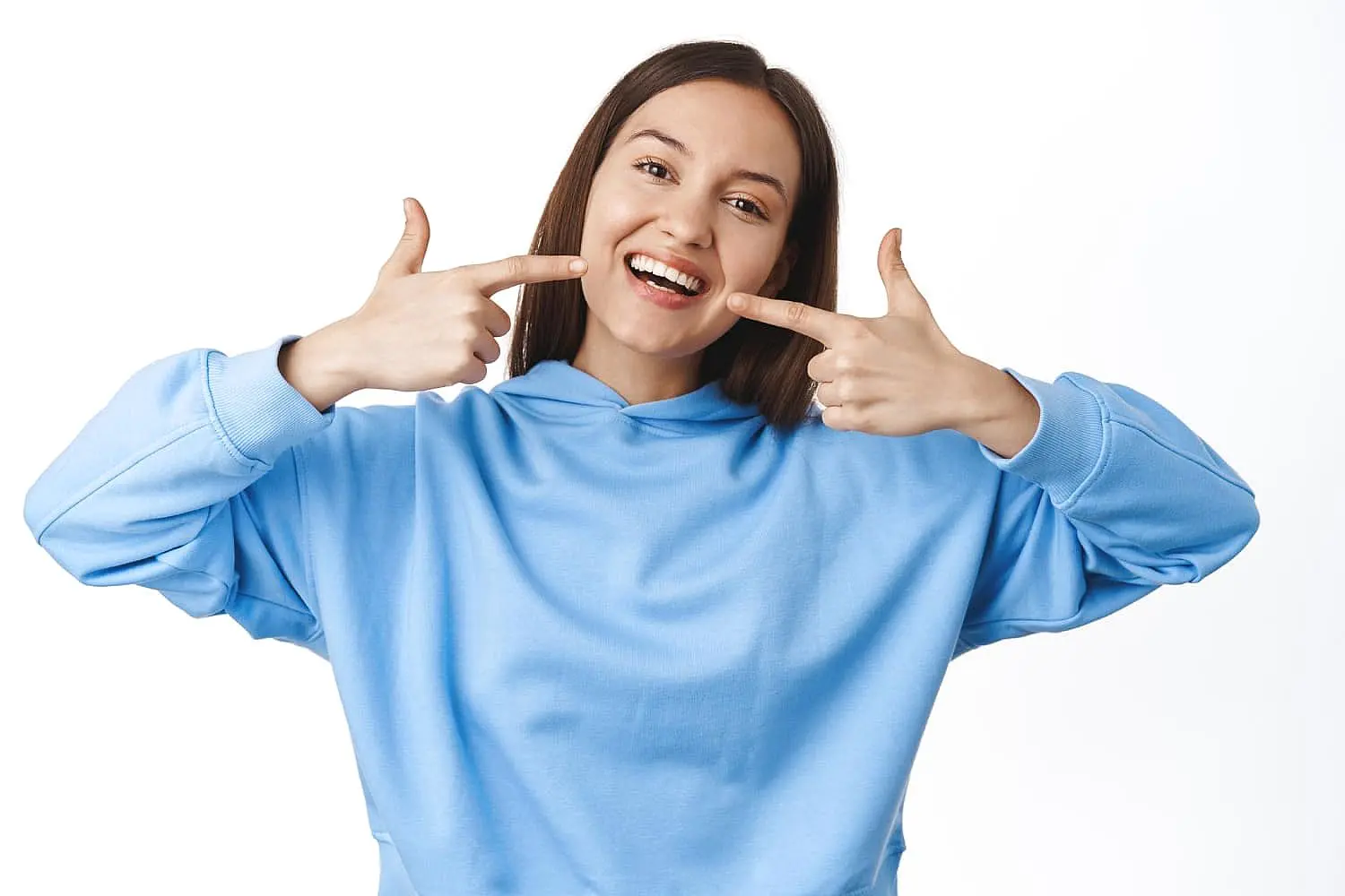 A woman in a blue hoodie smiles and points at her metal braces with both hands, proudly showcasing her orthodontic treatment at Smile City Orthodontics in Sunnyvale TX, against a white background.