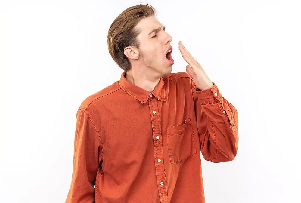 Standing before a plain white background, a man in an orange button-up shirt yawns and covers his mouth, resembling a typical mouth breather a reminder of why finding quality care at Smile City Orthodontics in Sunnyvale, TX can make all the difference.