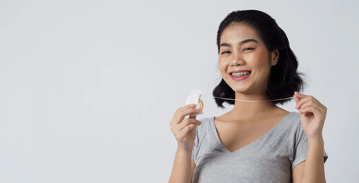 At Smile City Orthodontics in Sunnyvale TX, a patient with metal braces smiles while demonstrating proper flossing technique against a plain white background.