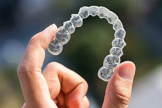 A hand displays a clear Invisalign aligner with a blurred background, showcasing Smile City Orthodontics in Sunnyvale, TX.