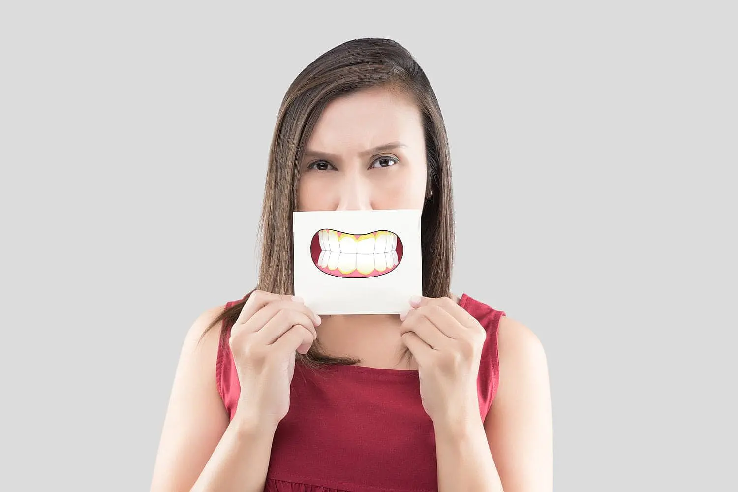 At Smile City Orthodontics in Sunnyvale TX, a woman wearing a red sleeveless top stands against a plain grey background, holding a paper with a cartoon underbite smile in front of her mouth.