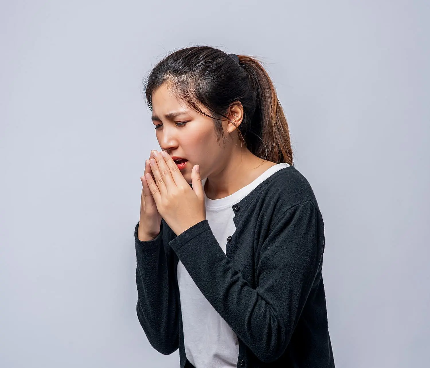 A woman with a ponytail, dressed in a white shirt and black cardigan, sneezes into her hands—possibly from mouth breathing—in front of a plain light gray background, highlighting concerns Smile City Orthodontics in Sunnyvale, TX can address.