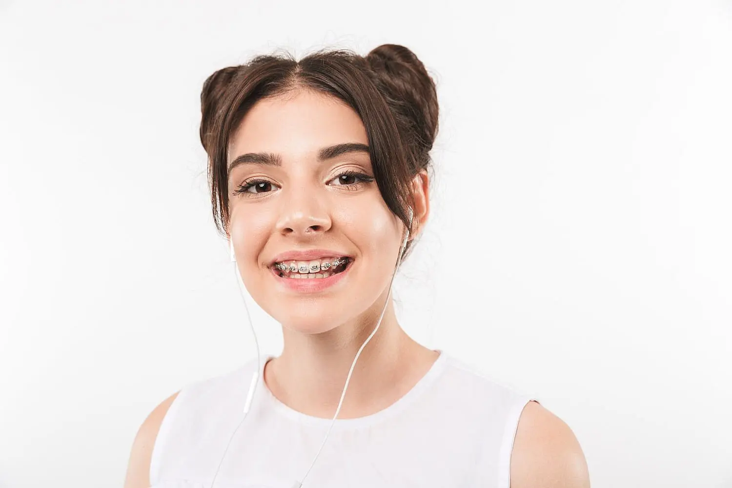 A young woman with dark hair in two buns, wearing metal braces and white earphones, smiles at the camera in a photo for Smile City Orthodontics in Sunnyvale, TX.