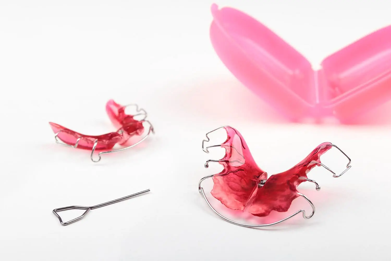 Displayed on a white background are two red orthodontic retainers essential tools used by a Children’s Orthodontist at Smile City Orthodontics in Sunnyvale, TX alongside a pink plastic storage case. These retainers, often used alongside Functional Appliances, help guide proper dental development in growing children.