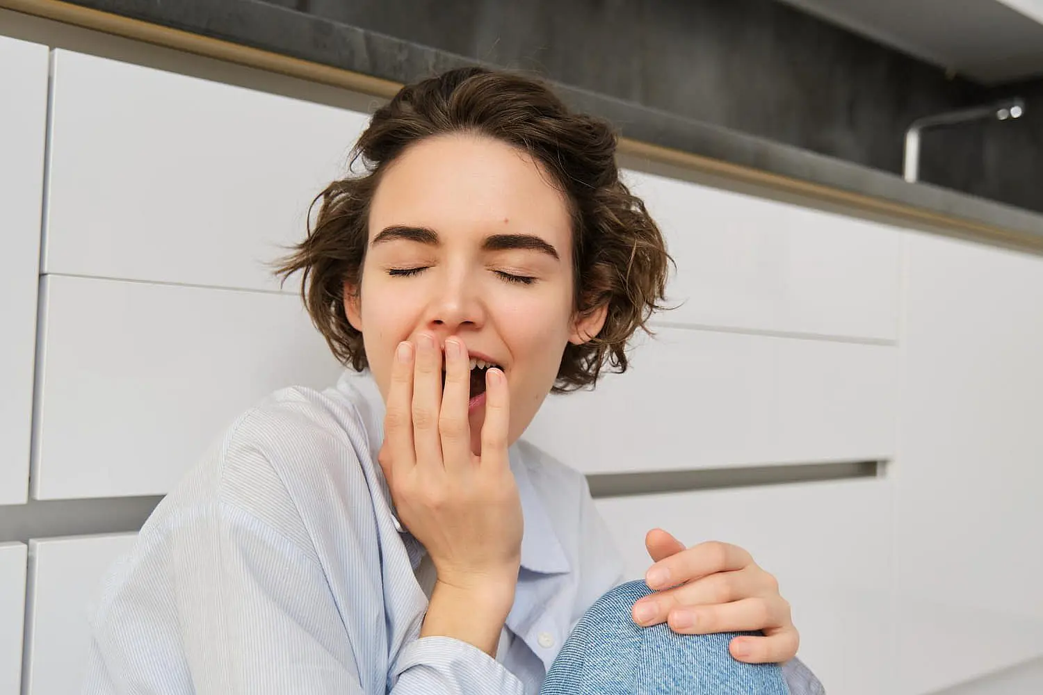 At Smile City Orthodontics in Sunnyvale TX, a person with short brown hair sits on the floor in front of white cabinets, covering their mouth with one hand as they yawn tiredly.