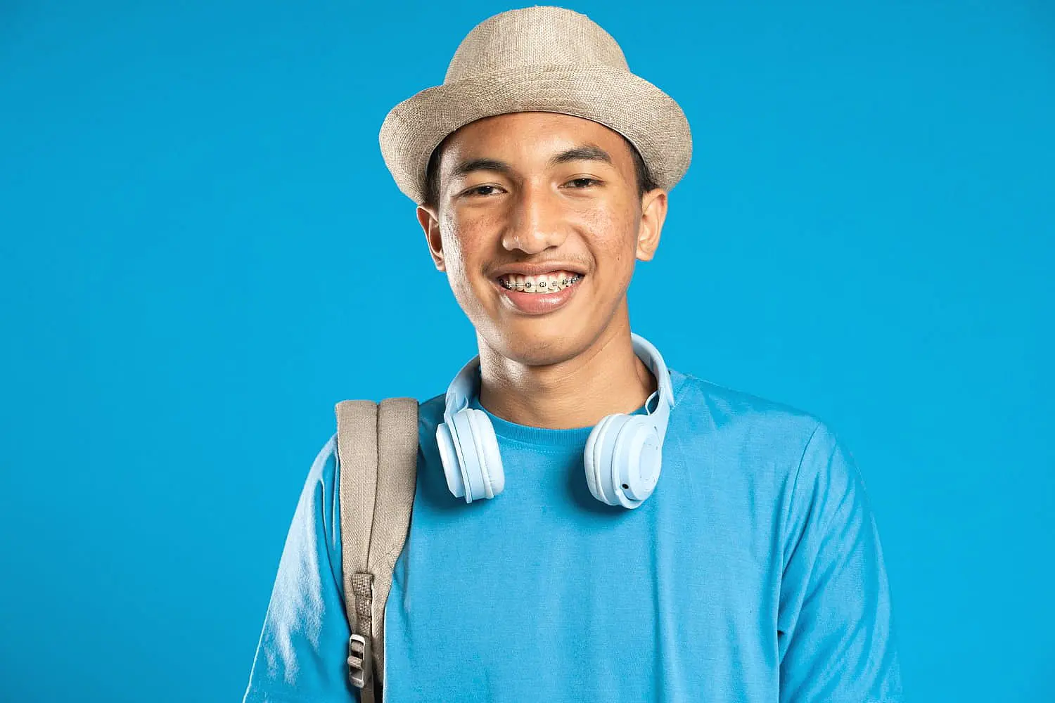 A young person in Sunnyvale, TX is pictured with an underbite, smiling with braces while wearing a hat, blue shirt, headphones around their neck, and a backpack strap over one shoulder against a blue background—representing Smile City Orthodontics.