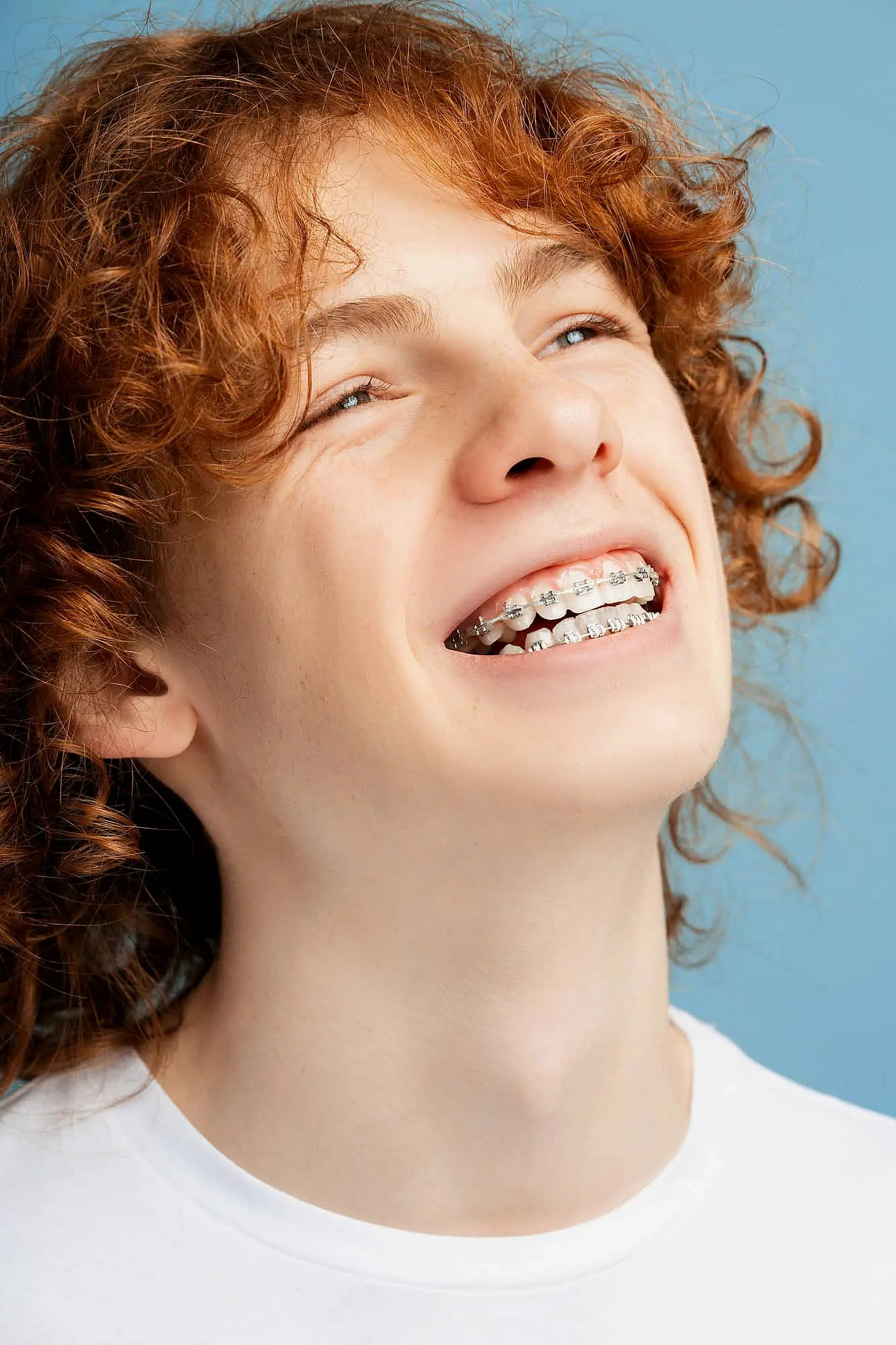 A teenager with curly red hair smiles, displaying metal braces against a blue background—demonstrating the confidence patients gain from treatments at Smile City Orthodontics in Sunnyvale, TX.