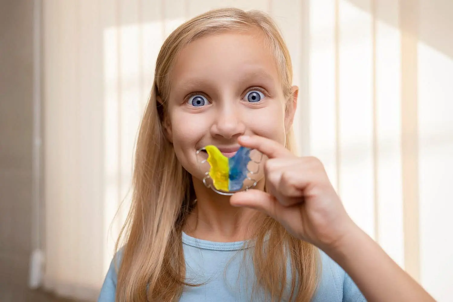 At Smile City Orthodontics in Sunnyvale, TX, a girl with long blonde hair holds a vibrant retainers featuring blue and yellow stripes, highlighting the creative options offered by their Children’s Orthodontist.