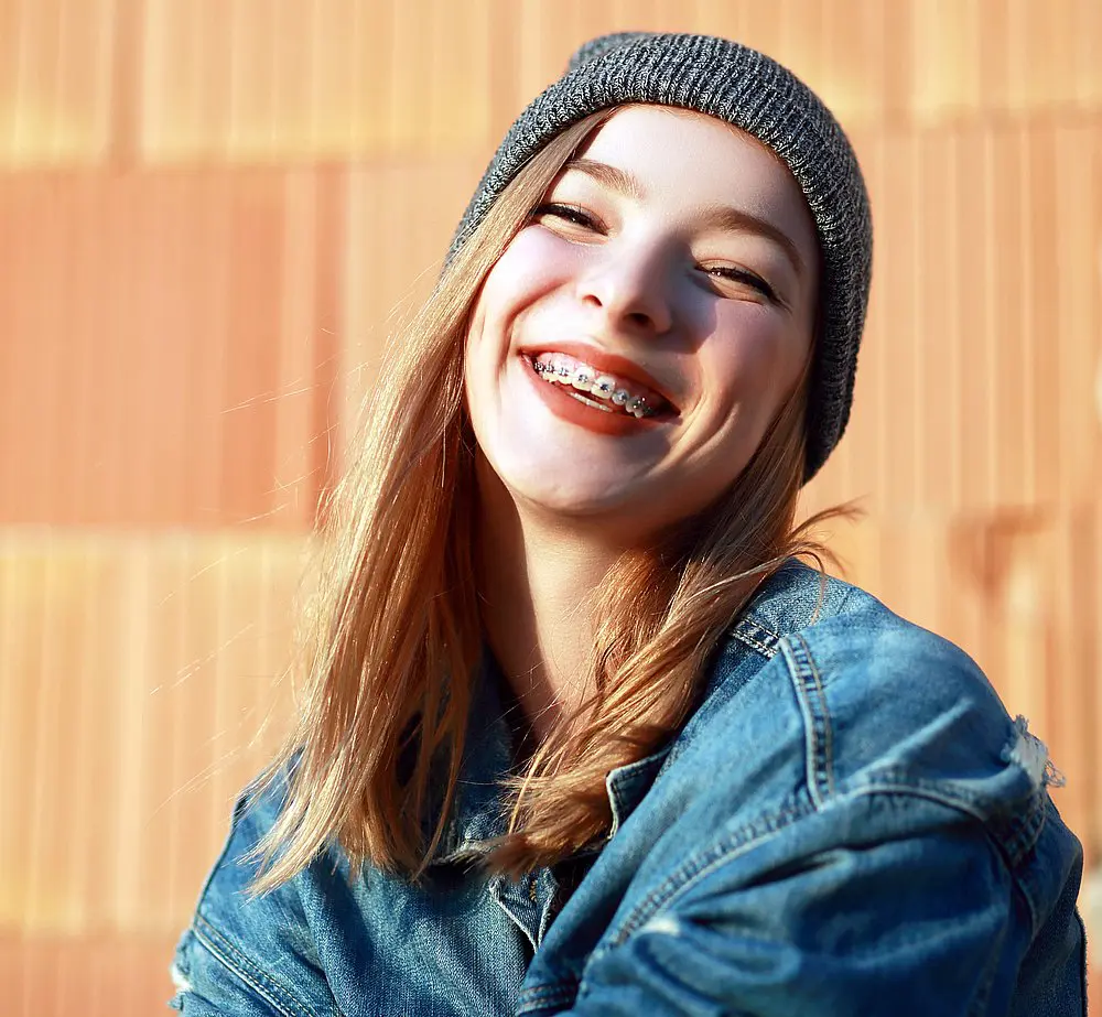 Highlighting the Invisalign vs. braces decision many locals face, Smile City Orthodontics in Sunnyvale, TX features a young woman in a gray beanie and denim jacket, smiling confidently and showing her braces against a peach-colored wall.