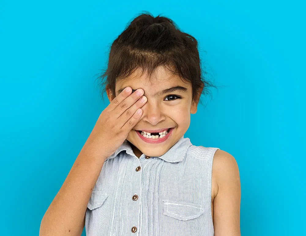 A young girl smiles and playfully covers one eye against a vibrant blue background an ideal image for Smile City Orthodontics in Sunnyvale, TX, highlighting happy, healthy smiles for a children’s orthodontist campaign.