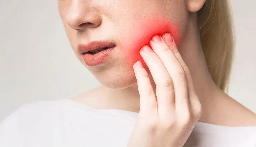 Someone touching their cheek with visible pain or swelling may be experiencing dental discomfort, and considering options like Invisalign vs. Braces at Smile City Orthodontics in Sunnyvale, TX for effective treatment.