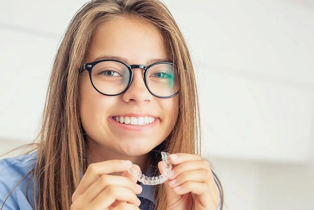A young woman with long hair and glasses smiles while holding clear aligners, highlighting the modern solutions offered by Smile City Orthodontics in Sunnyvale, TX, beyond traditional children's orthodontics.