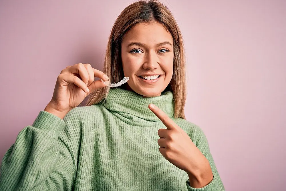 A woman in a green sweater stands before a pink background, smiling and pointing at a clear dental aligner she holds to highlight Invisalign Clear Aligners at Smile City Orthodontics in Sunnyvale, TX.