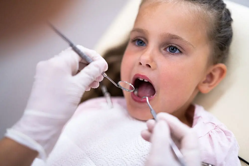 At Smile City Orthodontics in Sunnyvale TX, a dentist uses dental tools to check a young girl’s teeth for signs of tongue thrust as she sits in the chair.