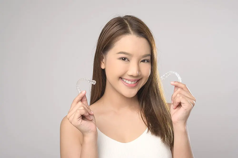 A woman with straight brown hair smiles and holds clear dental aligners against a plain background, showcasing the benefits of Invisalign at Smile City Orthodontics in Sunnyvale, TX—ideal for those exploring Invisalign insurance options.