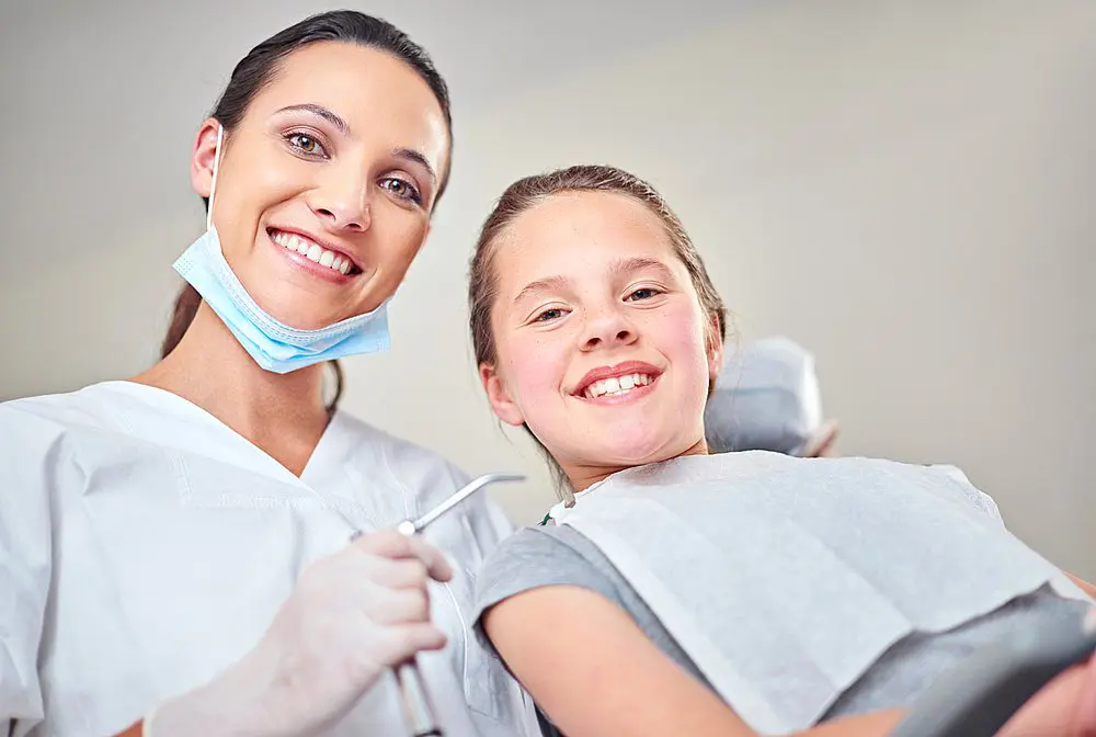 At Smile City Orthodontics in Sunnyvale TX, a children’s orthodontist wearing a mask and gloves stands beside a smiling young patient in the dental chair, both facing the camera.