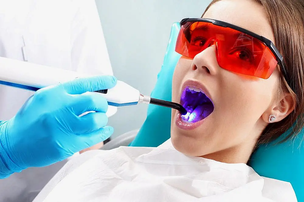 At Smile City Orthodontics in Sunnyvale TX, a dentist uses a curing light on a patient—equipped with red protective glasses and a bib—who is considering Invisalign vs Braces.