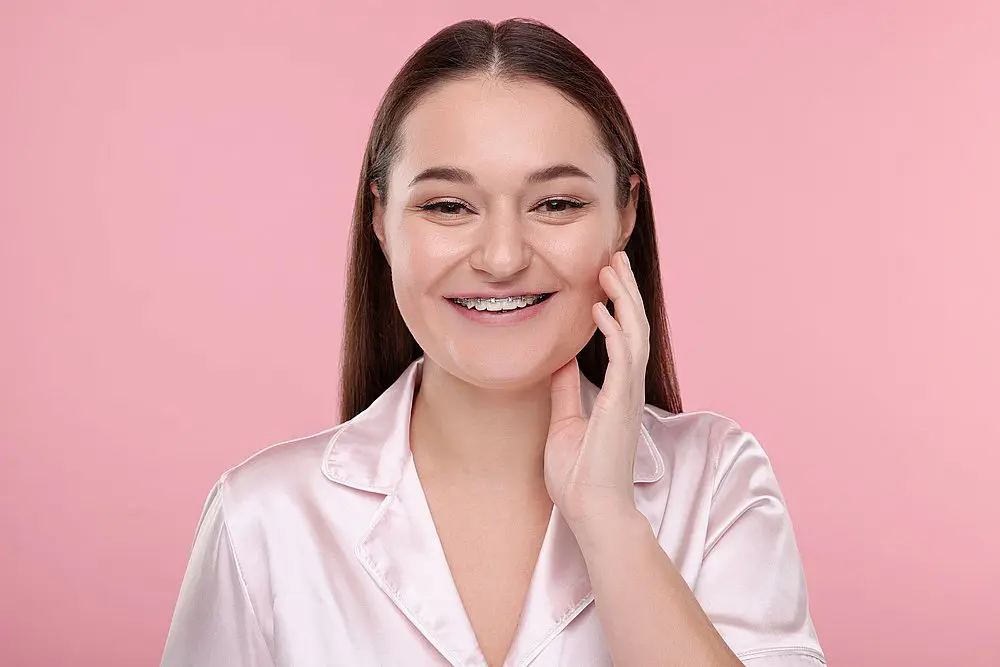 Showcasing the confidence that comes with adult braces, a woman with straight brown hair and braces smiles and touches her cheek in front of a pink background. This image highlights the transformative results provided by Smile City Orthodontics in Sunnyvale, TX.