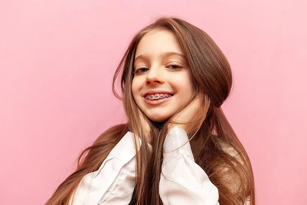 Showcasing the expertise of a children’s orthodontist at Smile City Orthodontics in Sunnyvale, TX, a young girl with long brown hair and traditional braces smiles brightly, holding her face with both hands against a pink background.
