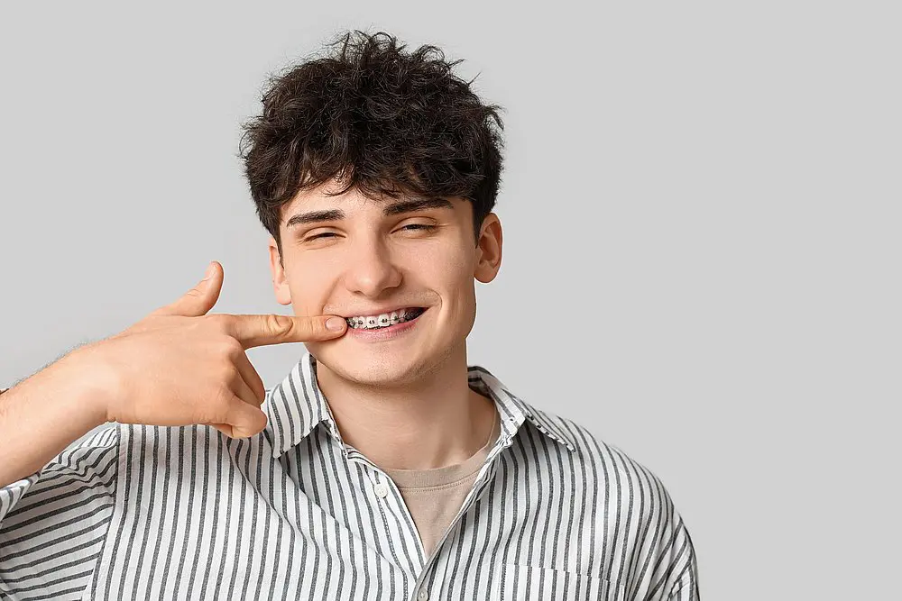A young man in a striped shirt stands against a plain gray background, smiling and pointing to his braces—highlighting the benefits of Adult Orthodontics at Smile City Orthodontics in Sunnyvale, TX.