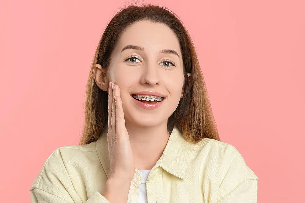 A young woman with long brown hair, wearing a yellow shirt and adult braces, smiles at the camera in front of a pink background at Smile City Orthodontics in Sunnyvale, TX.