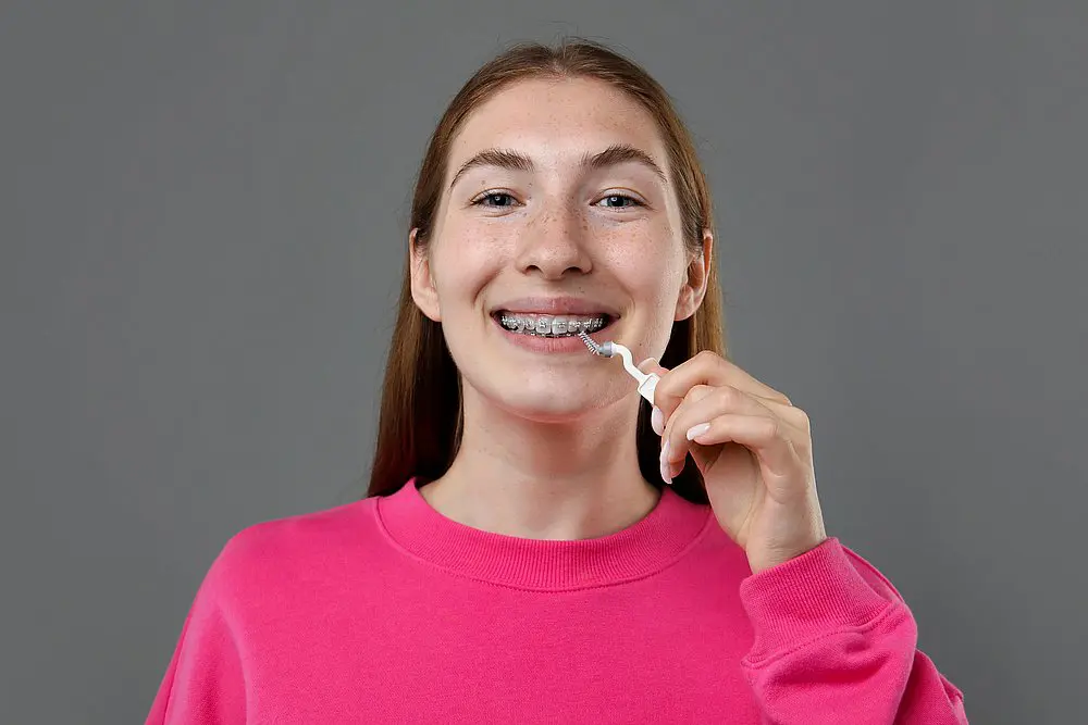 At Smile City Orthodontics in Sunnyvale TX, a young person with adult braces demonstrates proper oral care by using an interdental brush, dressed in a bright pink sweatshirt against a simple gray background.