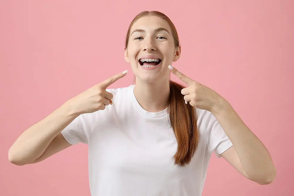 A young woman with long red hair wearing a white t-shirt smiles and points to her metal braces in a photo for Smile City Orthodontics in Sunnyvale TX, set against a pink background.