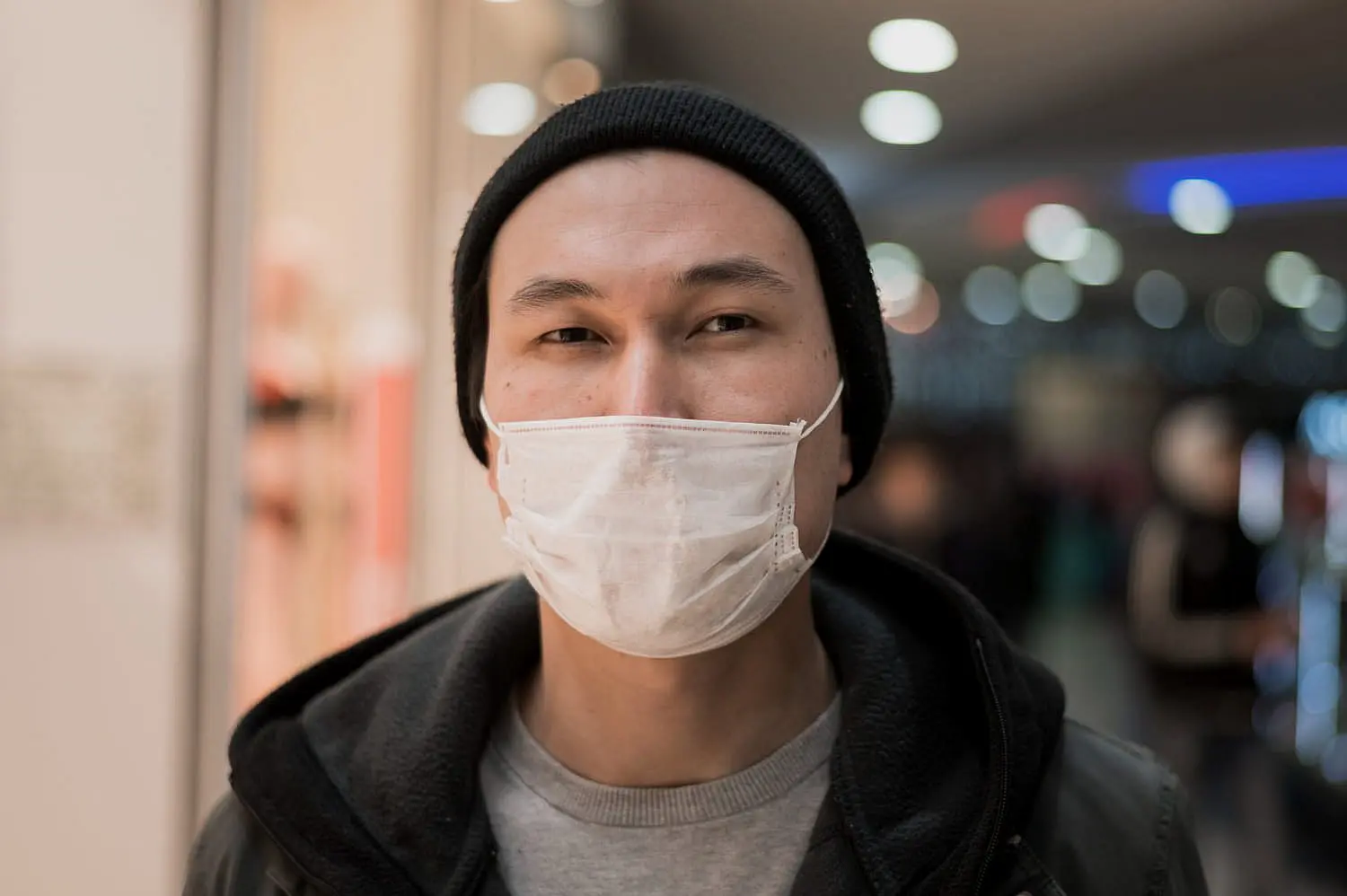 Indoors at Smile City Orthodontics in Sunnyvale TX, a person in a black beanie, white face mask—suggesting mouth breathing—and dark jacket stands before blurred lights.