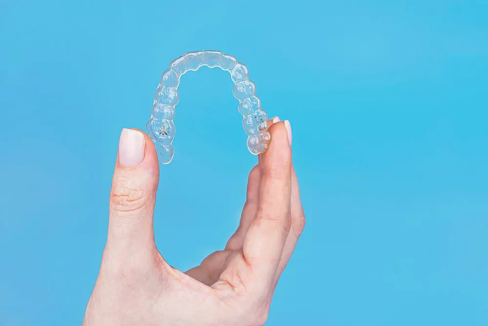 A hand holds a clear dental aligner against a plain blue background, showcasing options like Invisalign cost at Smile City Orthodontics in Sunnyvale, TX.
