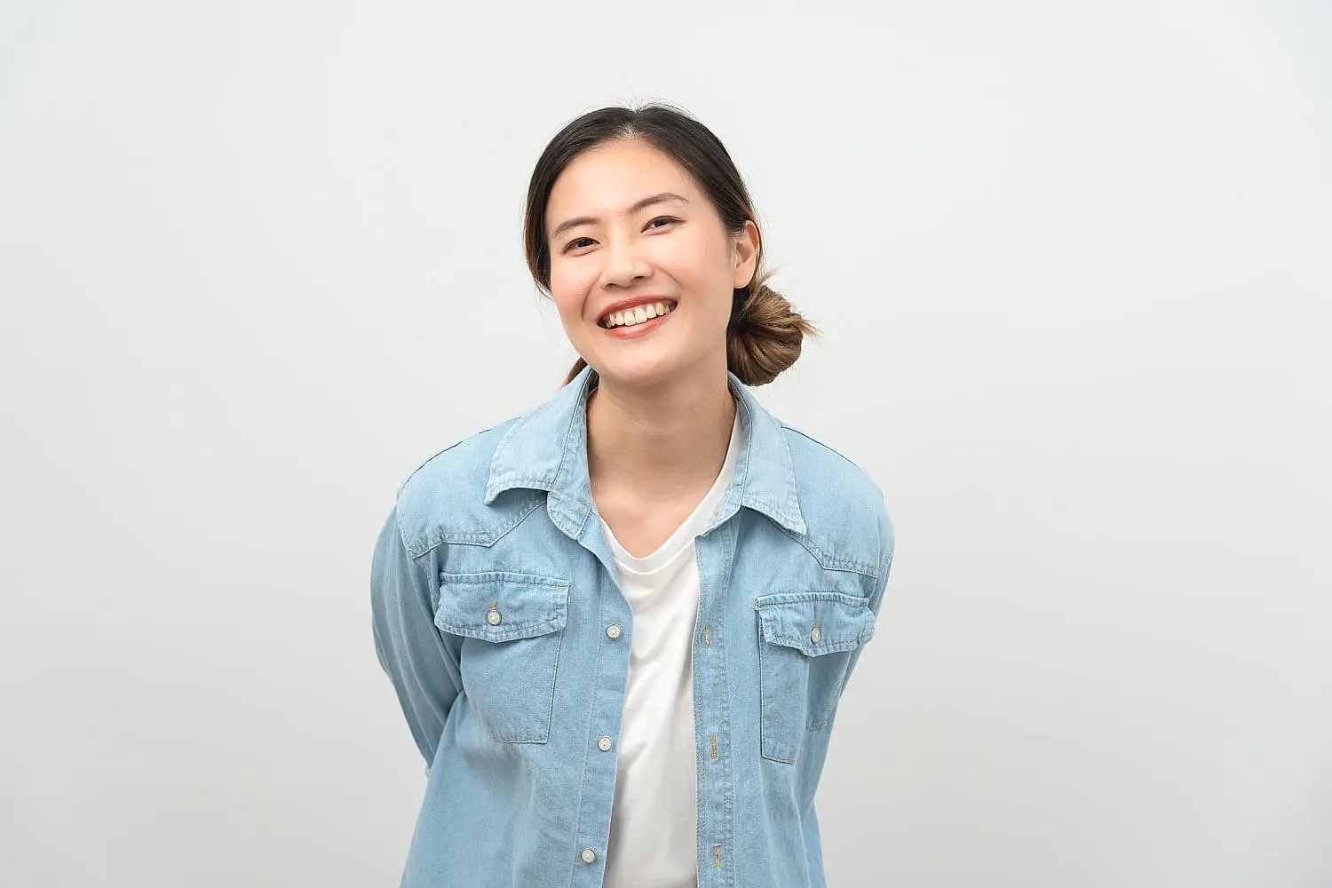 A woman in a light denim shirt and white top stands against a plain background, confidently smiling with her hands behind her back—demonstrating the benefits of Invisalign from Smile City Orthodontics in Sunnyvale, TX.