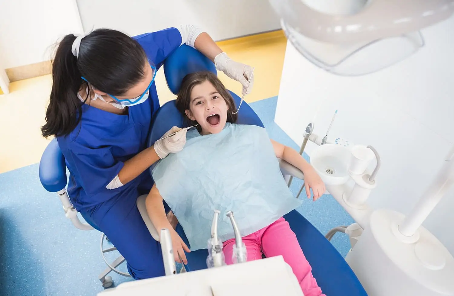 At Smile City Orthodontics in Sunnyvale TX, a dentist in blue scrubs examines a young girl’s mouth as she sits with a bib in a well-lit clinic equipped with orthodontic appliances.