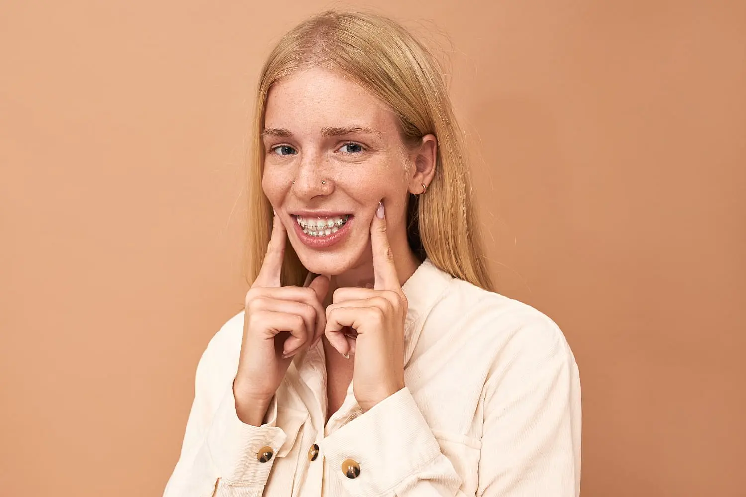 A young woman with long blonde hair stands against a beige background, smiling and pointing to her clear braces—showcasing her confident results from Smile City Orthodontics in Sunnyvale, TX.