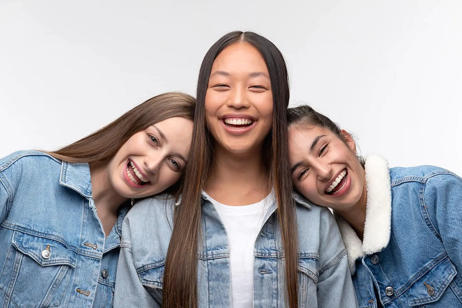Three young women in denim jackets stand close together, smiling and laughing against a plain white background—the kind of confidence you can expect at Smile City Orthodontics in Sunnyvale, TX.