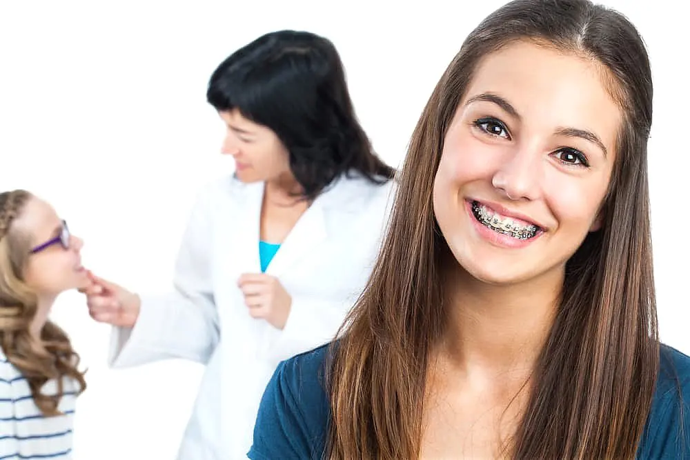 Smile City Orthodontics A teenage girl with braces smiles confidently at the camera while a dentist attends to another patient, showcasing the expert care and affordable braces cost provided by Smile City Orthodontics in Sunnyvale, TX.
