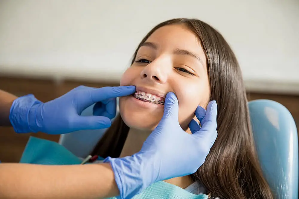 Smile City Orthodontics At Smile City Orthodontics in Sunnyvale TX, a dentist wearing blue gloves examines a smiling young girl’s braces and discusses the cost of braces.