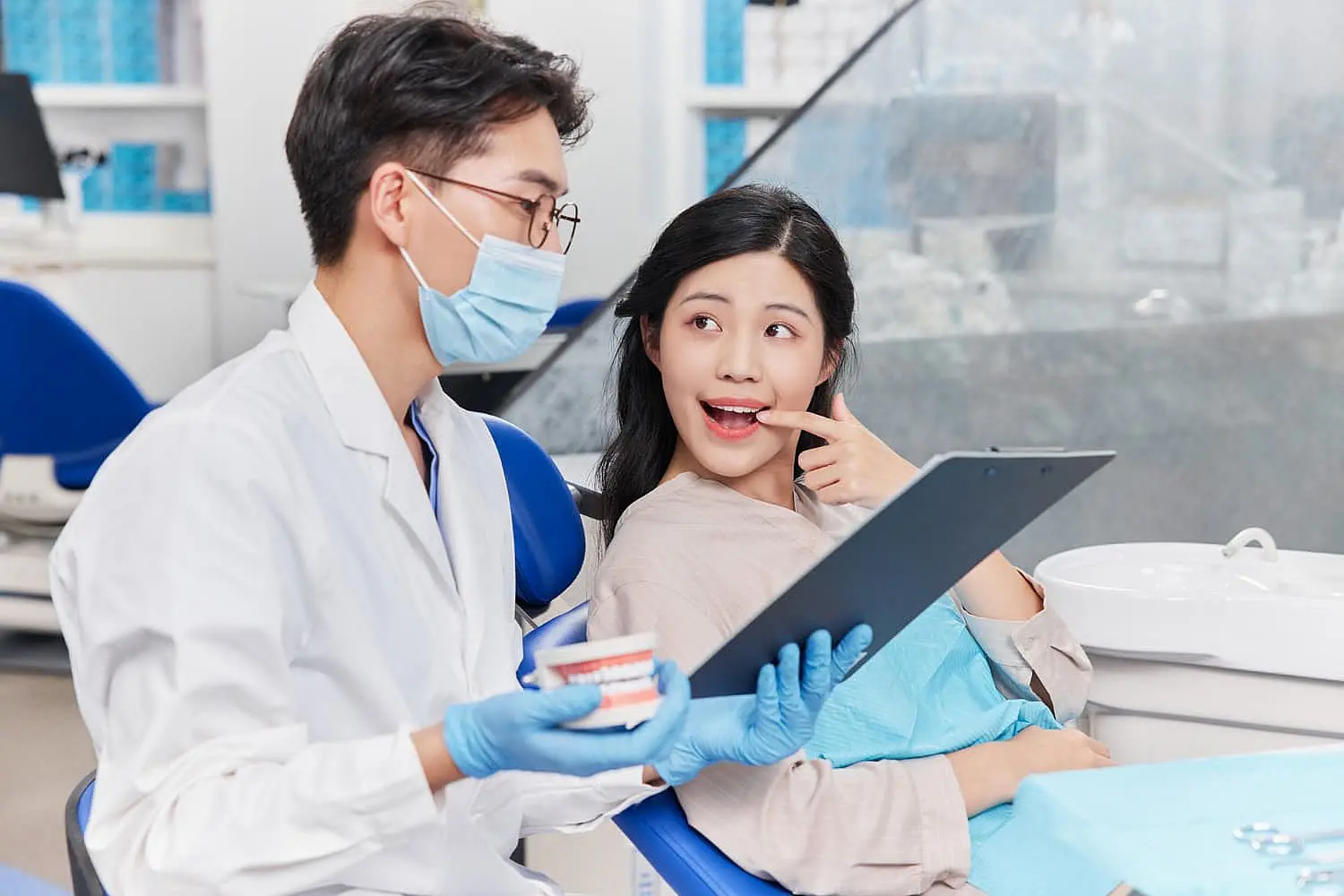 At Smile City Orthodontics in Sunnyvale TX, a dentist wearing a mask and gloves uses a dental model to explain jaw surgery to a female patient as she points to her teeth.