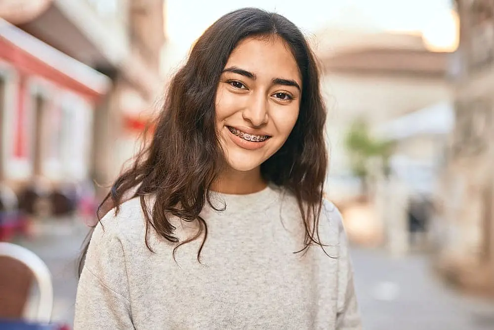 A young person in Sunnyvale TX, with long dark hair and metal braces from Smile City Orthodontics smiles outdoors on a city street, dressed in a light gray sweater.