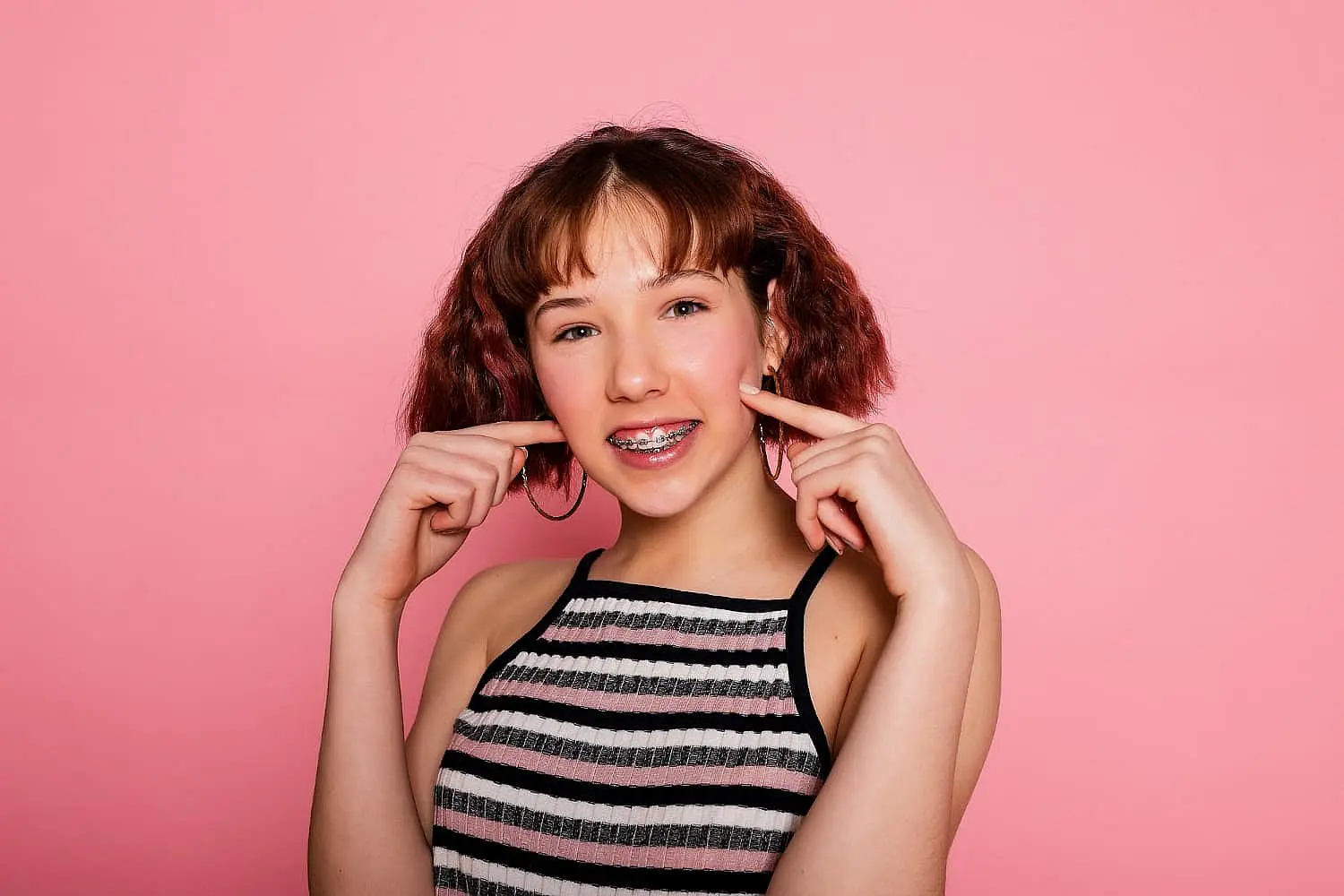 Against a plain pink background, a teenager with short brown hair and braces smiles and points to her cheeks, embodying the essence of Invisalign vs Braces at Smile City Orthodontics in Sunnyvale, TX.