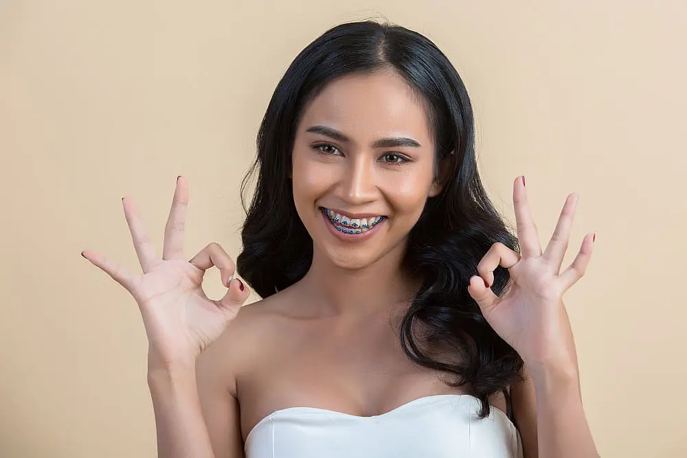 Confident after getting metal adult braces at Smile City Orthodontics in Sunnyvale TX, a woman with long black hair smiles and flashes "OK" hand signs against a plain beige background.