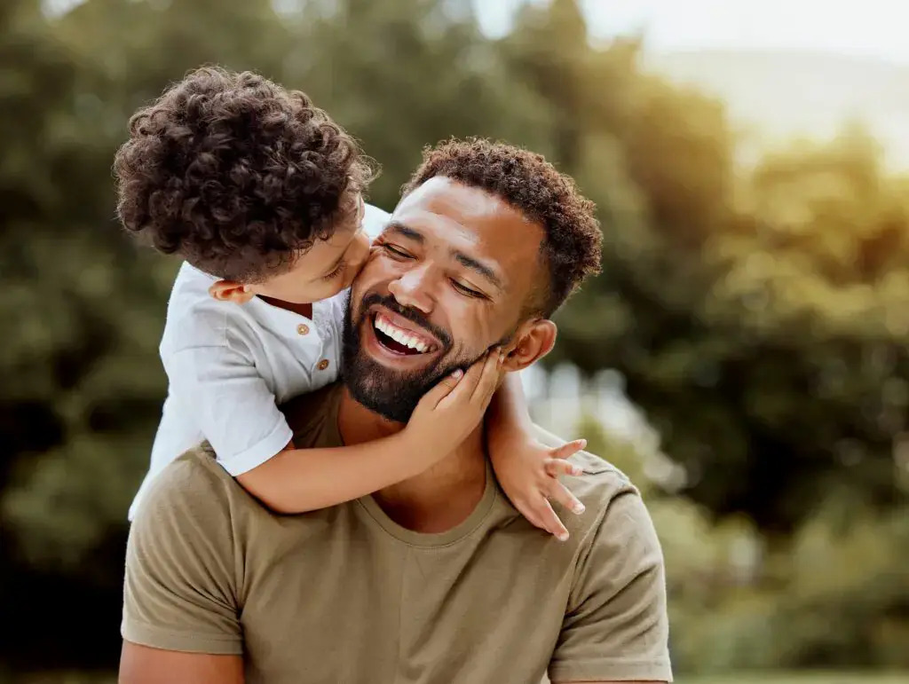 At Smile City Orthodontics in Sunnyvale TX, a smiling man is hugged and kissed on the cheek by a young child outdoors, with trees softly blurred in the background. At Smile City Orthodontics in Sunnyvale TX, a smiling man is hugged and kissed on the cheek by a young child outdoors, with trees softly blurred in the background.