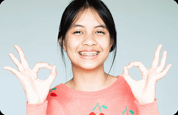 A smiling girl with metal braces and a pink sweater makes an "OK" gesture against a plain light background, representing Smile City Orthodontics in Sunnyvale, TX.