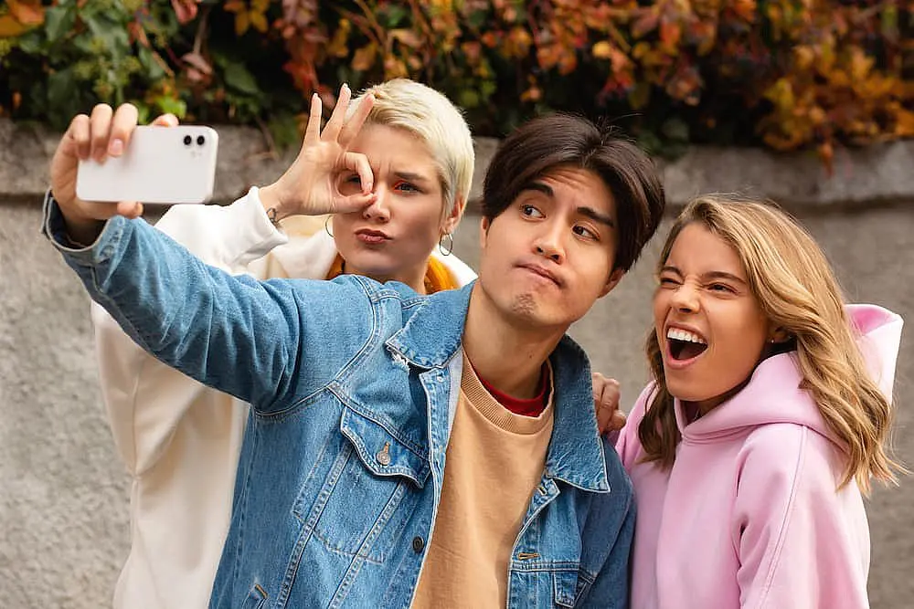 Three young adults pose for a selfie outdoors, with one holding the phone, another giving an “OK” sign, and the third showing off their smile—an ideal scene for Smile City Orthodontics in Sunnyvale, TX to highlight various types of braces.