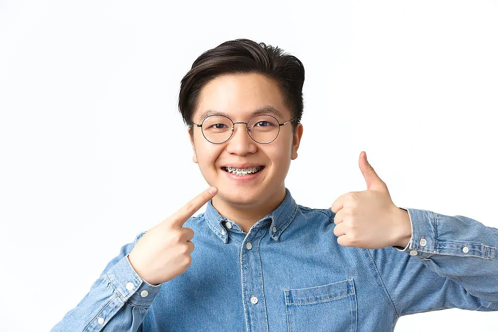 A person wearing glasses and braces confidently smiles, points to their braces, and gives a thumbs up, highlighting their positive experience with adult braces at Smile City Orthodontics in Sunnyvale TX, against a white background.