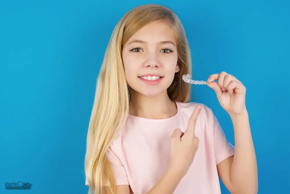 Smiling young girl with long blonde hair holding a clear aligner after understanding Does Insurance Cover Invisalign in Sunnyvale, TX in front of a blue background.