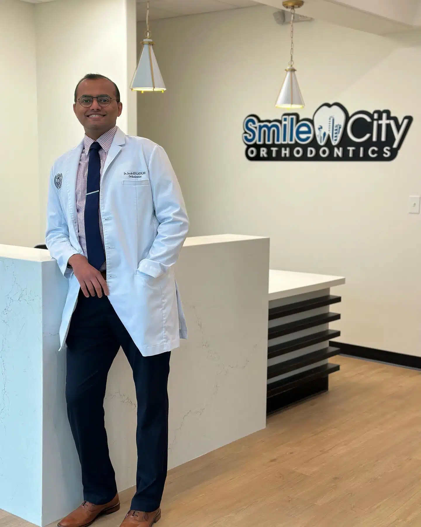At Smile City Orthodontics in Mesquite TX, Dr. Jacob an orthodontics expert in a white lab coat, stands by the reception desk with the clinic’s sign visible behind him.