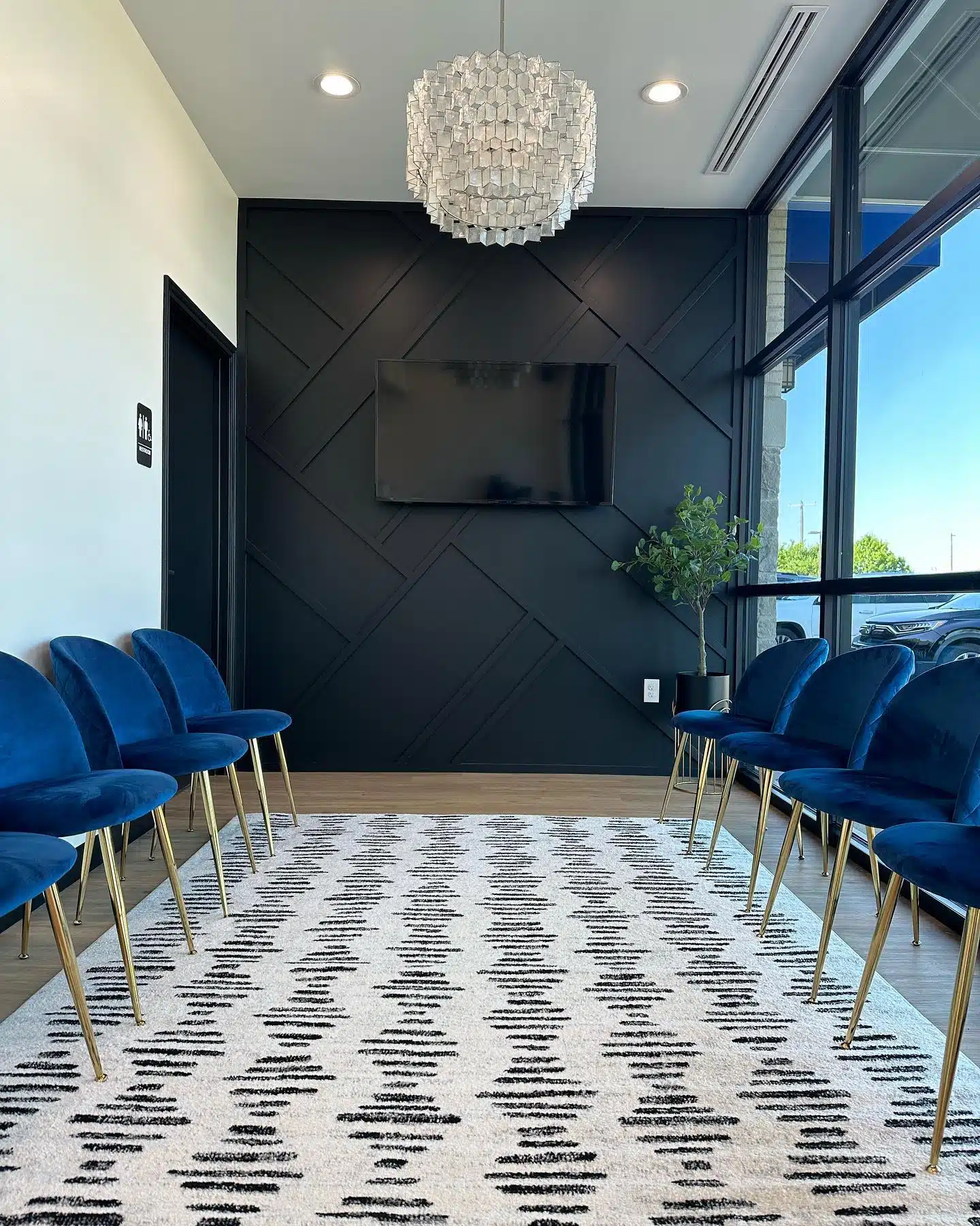 At Smile City Orthodontics in Sunnyvale TX, the modern waiting room features blue velvet chairs, a geometric rug, and a sleek black accent wall with dental art focused on overbite care. Large windows fill the space with natural light beneath a striking hanging light fixture.