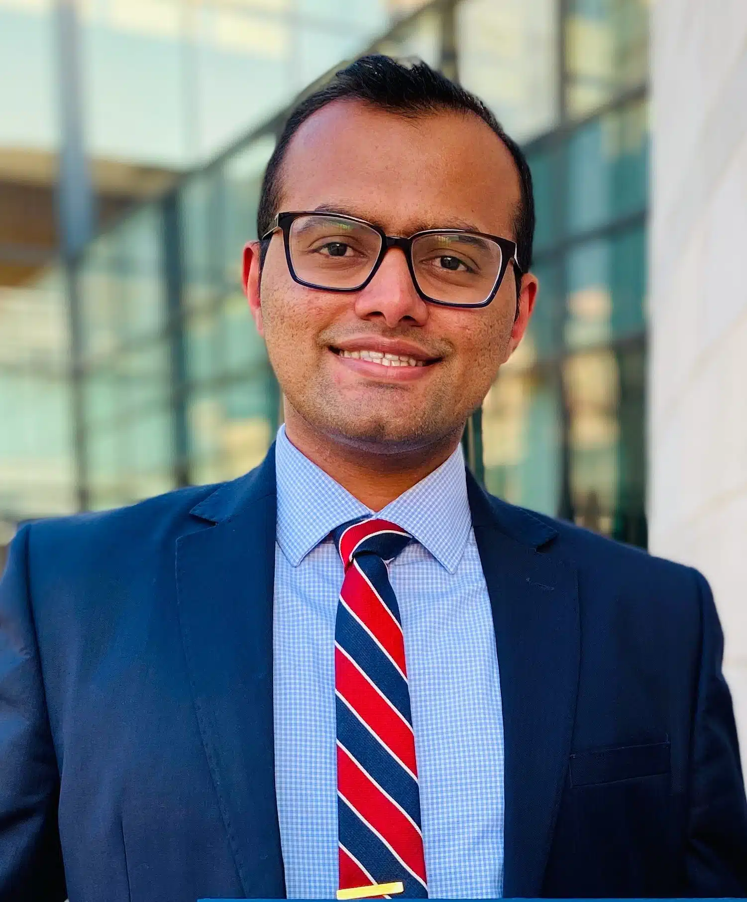 At Smile City Orthodontics in Sunnyvale TX, Dr. Jacob BDS, MS an orthodontic expert smiles at the camera outside a modern glass building, dressed in glasses, a dark suit, blue checked shirt, and red striped tie.