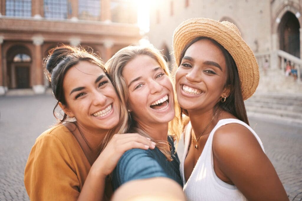 Three young women smile and take a selfie outdoors in a sunlit, urban setting with historic buildings, close to Smile City Orthodontics in Sunnyvale, TX.